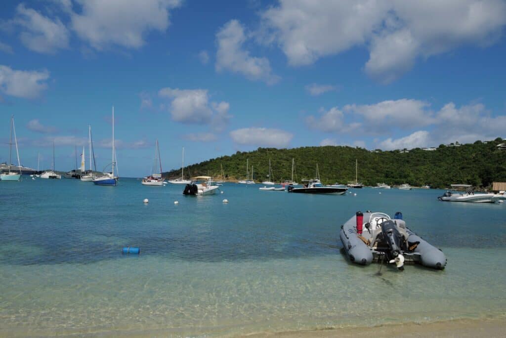 A grey inflatable motorboat anchored in shallow water with sailboats moored in the bay beyond a green hill near Cruz Bay on St. John, USVI.