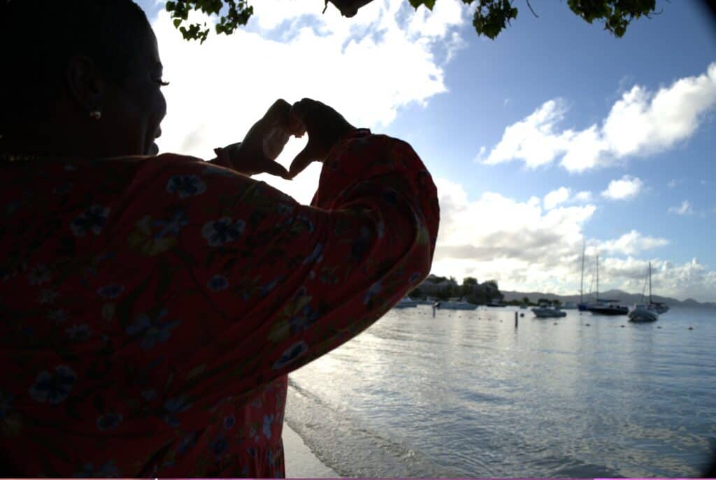 A person in a patterned dress forms a heart shape with their hands against a backdrop of the ocean and sailboats at Cruz Bay on St. John, USVI.