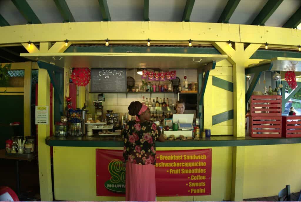 A woman standing at a yellow walk-up cafe counter at Sib's Daily Dose on St. Thomas, USVI, while a staff member works at a register; a menu board hangs above the service area.