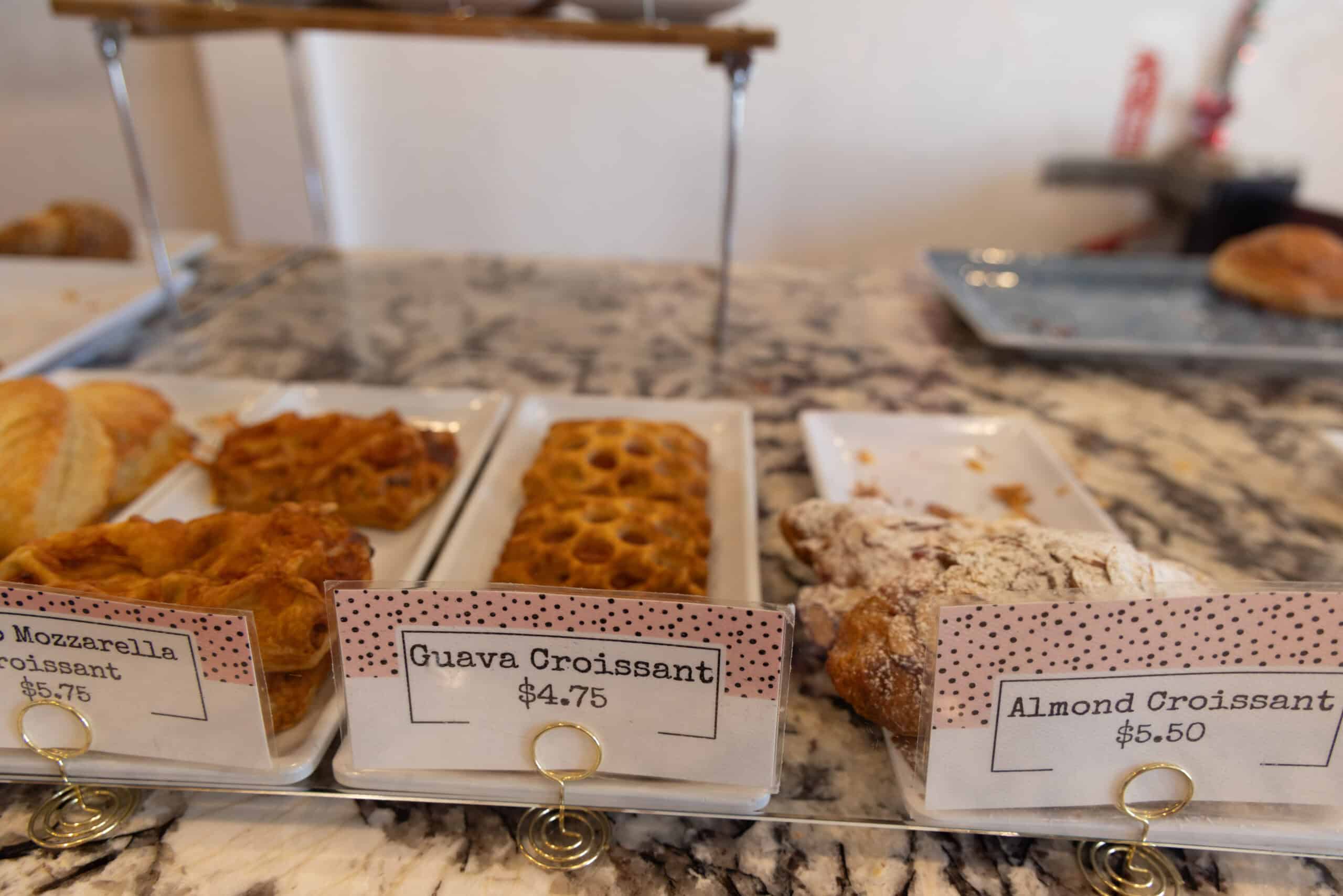 Three rectangular platters of pastries on a marble counter, with signs labeling them as mozzarella croissant, guava croissant, and almond croissant with prices. Stir It Up Coffee Shop & Bakery on St. Thomas.