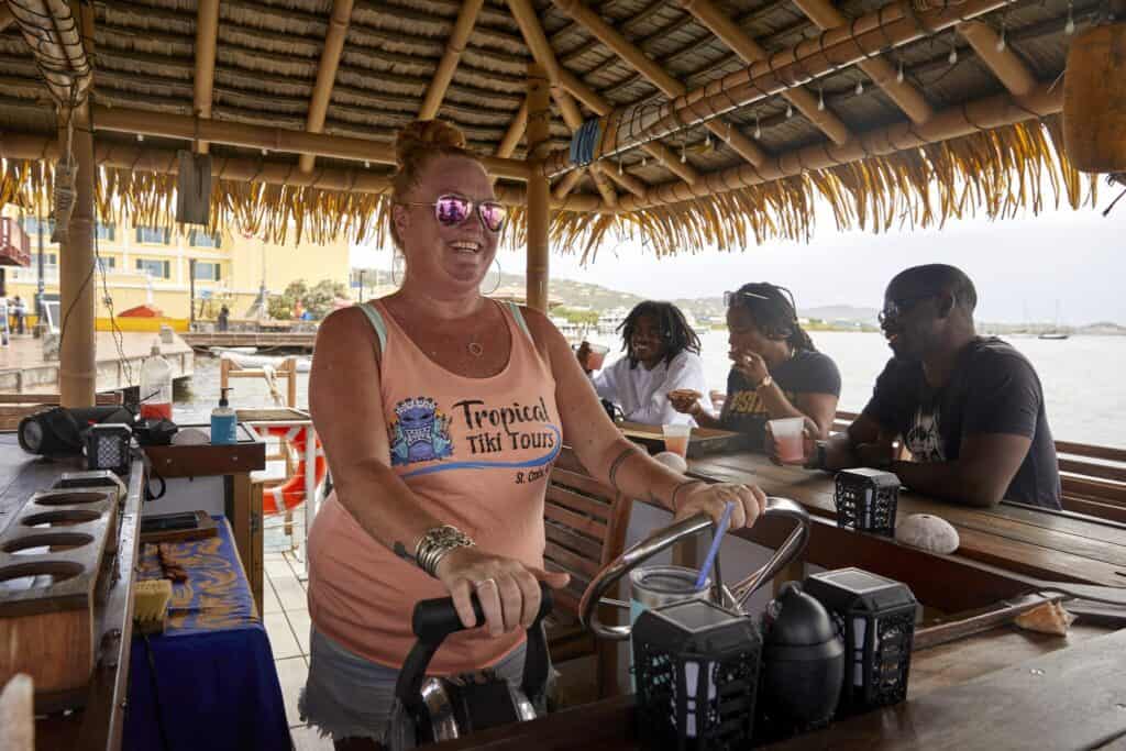 A woman wearing sunglasses and a Tropical Tiki Tours tank top stands at the helm of a tiki bar boat with three customers seated at the bar behind her in the waters off St. Croix, USVI.